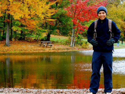 A person stands by a lake surrounded by colorful autumn trees, wearing a backpack and smiling.