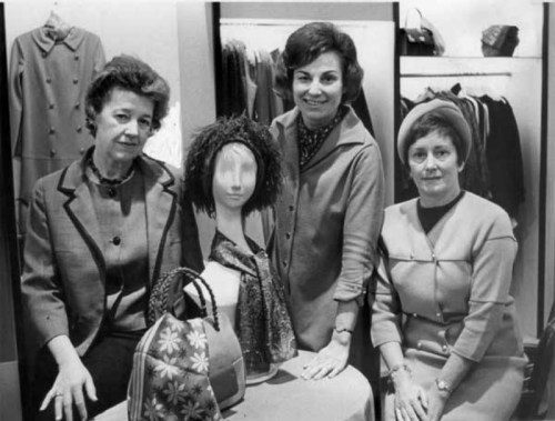 Three women pose with a mannequin wearing a wig, surrounded by clothing and accessories in a boutique setting.