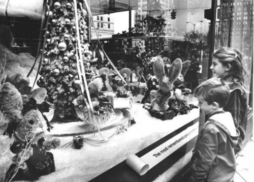 Two children admire a festive holiday window display featuring a decorated tree and plush animals.