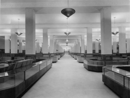 A spacious, empty bank interior with tall columns, glass display cases, and vintage light fixtures.