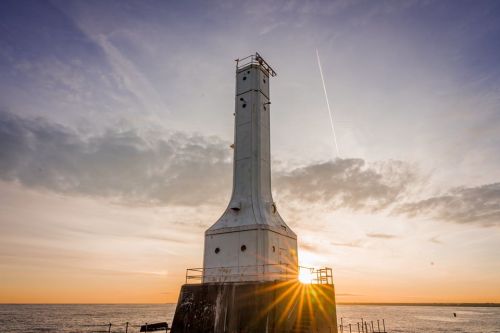 A tall, white lighthouse silhouette against a sunset sky, with sun rays shining through the structure over the water.