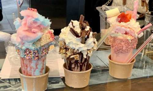 Three colorful milkshakes topped with whipped cream, candy, and cotton candy, served in paper cups.