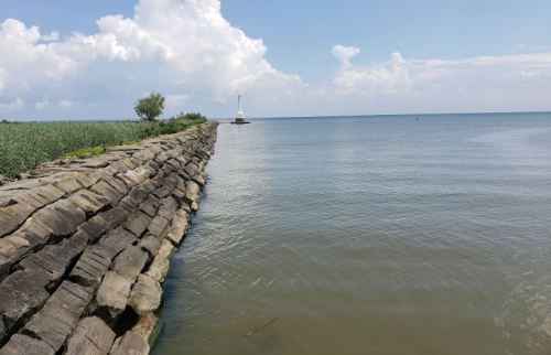 A rocky shoreline extends into calm water, with a lighthouse visible in the distance under a blue sky with clouds.