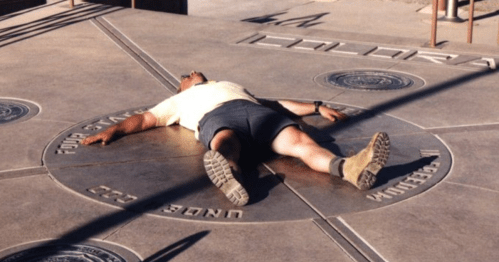 A person lies on the ground, arms outstretched, on a circular concrete design in a sunny outdoor setting.