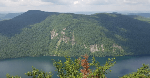 A lush green mountain rises above a calm lake, with distant hills under a partly cloudy sky.