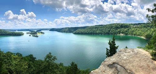 A panoramic view of a serene lake surrounded by lush green hills and a bright blue sky with fluffy clouds.