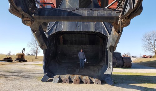 A person stands inside the massive bucket of a large excavator, showcasing its size against a clear blue sky.