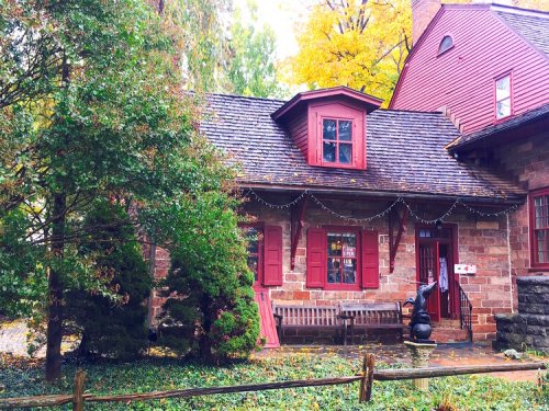 A charming stone house with red accents, surrounded by greenery and autumn leaves, featuring a bench and festive lights.