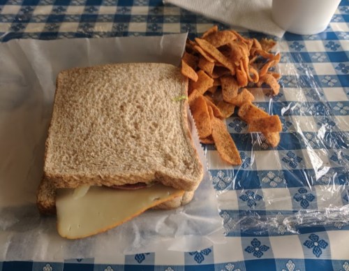 A sandwich on whole grain bread with cheese, next to a pile of crunchy snack chips, on a blue checkered tablecloth.