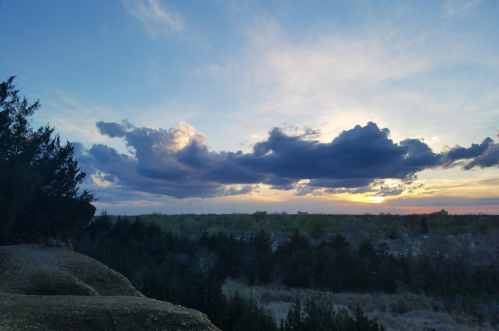 A scenic view of a sunset with colorful clouds over a grassy landscape and distant trees.