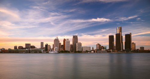 A skyline view of Detroit at sunset, featuring tall buildings and a calm river in the foreground.