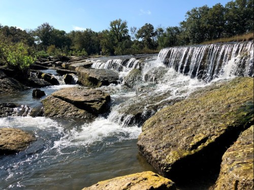 A serene waterfall cascades over rocky ledges, surrounded by lush greenery and a clear blue sky.