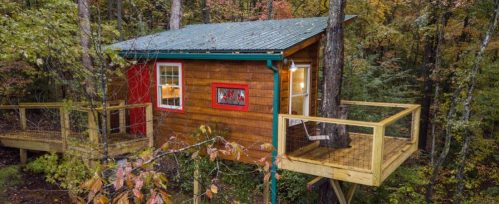 A cozy treehouse with a wooden exterior, surrounded by trees and featuring a small deck and colorful window accents.