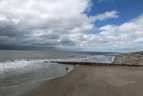 A beach scene with a person walking along the shore, cloudy skies, and gentle waves lapping at the sand.