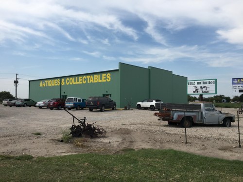 A green building with "Antiques & Collectables" sign, surrounded by parked cars and a gravel lot.
