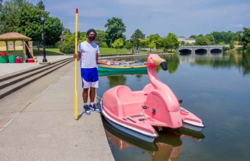 A person stands by a pink flamingo paddle boat on a calm lake, with trees and a bridge in the background.