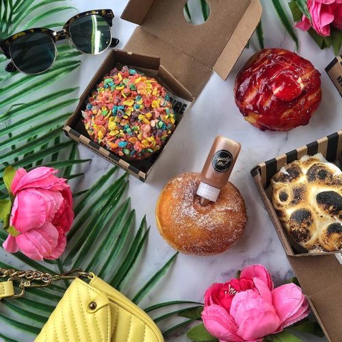 Colorful donuts in boxes surrounded by tropical leaves, pink flowers, sunglasses, and a bottle of syrup.