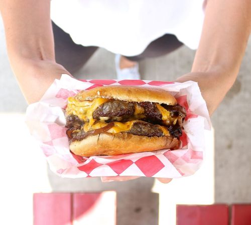 A person holds a cheeseburger with melted cheese and grilled meat, wrapped in red and white checkered paper.