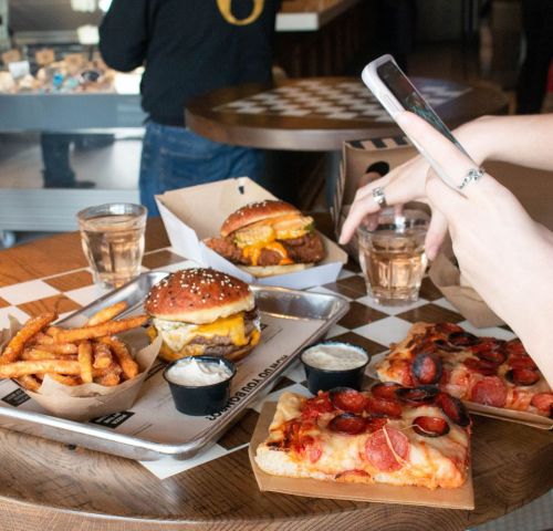 A table with a burger, pizza, fries, and drinks, with a hand holding a phone taking a photo.