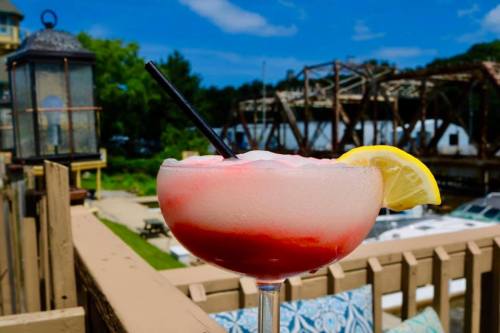 A colorful frozen cocktail with a lemon slice, set against a sunny waterfront backdrop.