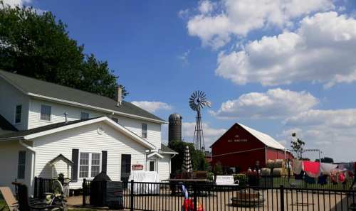A farm scene featuring a white house, red barn, windmill, and clothes drying outside under a blue sky with clouds.