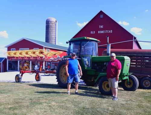 Two men stand beside a green tractor with a farming implement, in front of a red barn and silo on a sunny day.