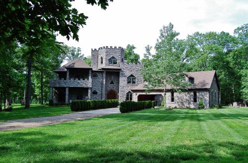 A large stone house with a turret, surrounded by trees and a manicured lawn on a sunny day.