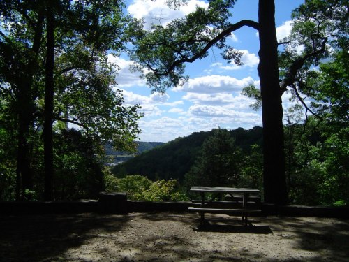 A scenic view of a valley with trees and a picnic table under a bright blue sky with fluffy clouds.