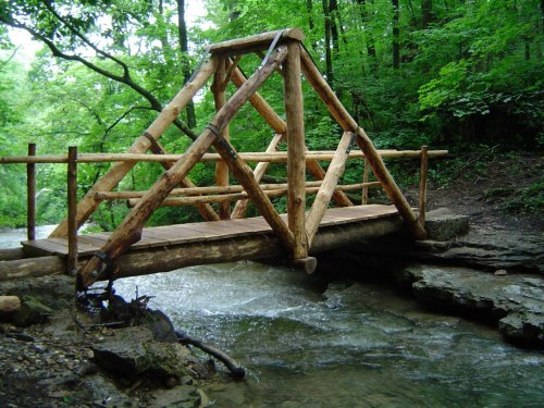 A wooden bridge with a triangular design spans a small stream, surrounded by lush green trees and foliage.