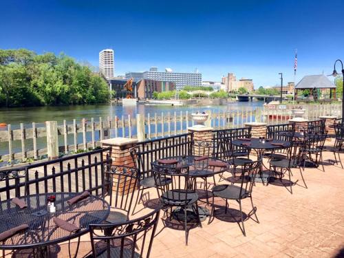 Outdoor seating area by a river, with tables and chairs, surrounded by greenery and city buildings in the background.