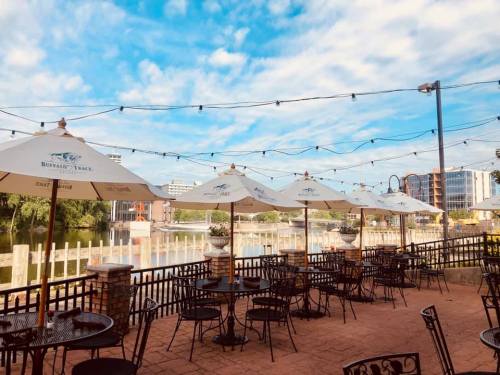Outdoor dining area with tables and umbrellas by a river, under a blue sky with scattered clouds and string lights.
