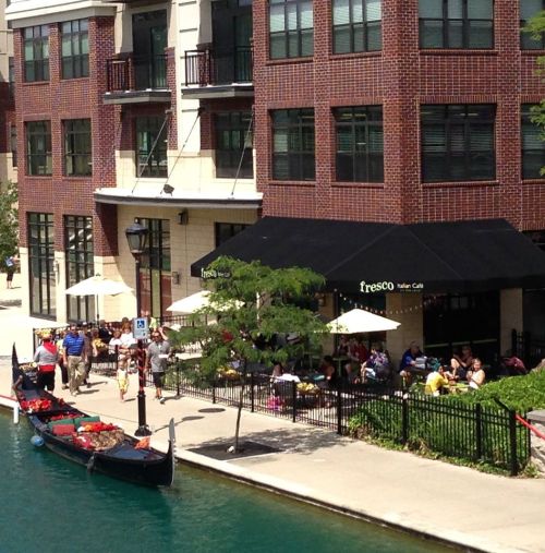 A canal-side Italian café with outdoor seating, surrounded by brick buildings and a gondola in the water.