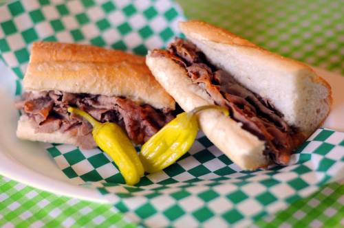 A close-up of a roast beef sandwich on a hoagie roll, served with two yellow peppers on a green checkered plate.