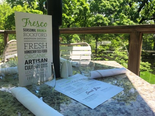 A table set for dining outdoors, featuring menus, glasses, and napkins, surrounded by greenery.