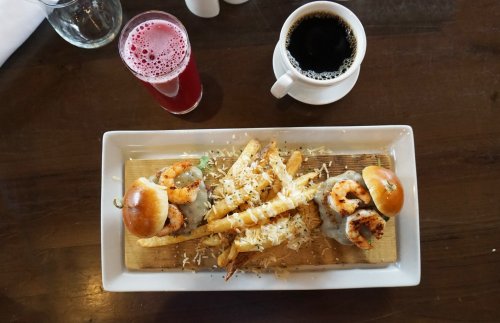 A wooden platter with two shrimp sliders, fries topped with cheese, a glass of pink drink, and a cup of black coffee.