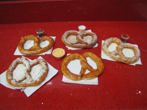 Five large pretzels on a red countertop, two with powdered sugar, served with dipping sauces.