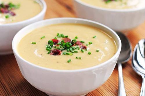 Creamy soup in a white bowl, topped with chopped chives and bits of bacon, on a wooden table.