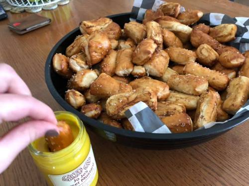 A hand dips a pretzel bite into a jar of mustard, with a large bowl of pretzel bites in the background.