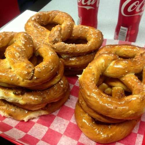 Two stacks of soft pretzels sprinkled with salt, accompanied by two cans of Coca-Cola on a red-checkered tray.
