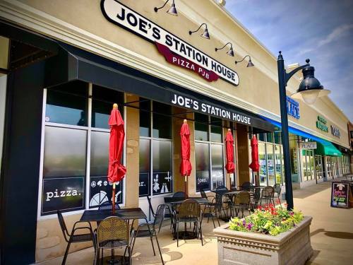 Exterior of Joe's Station House Pizza Pub, featuring outdoor seating with red umbrellas and a flower bed.
