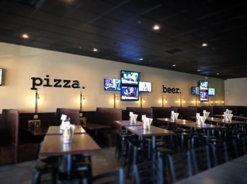 Interior of a restaurant with a wall displaying "pizza" and "beer," featuring multiple TVs and wooden tables.