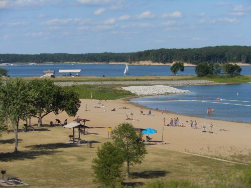 A sandy beach with people sunbathing and swimming, surrounded by water and trees under a blue sky with clouds.