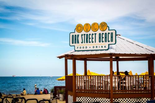 Sign for Oak Street Beach with a beachside gazebo and people enjoying the view of the water and yellow umbrellas.
