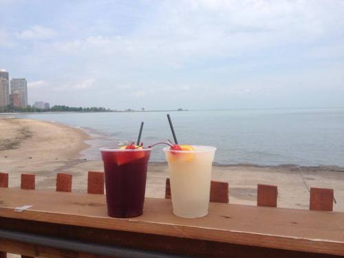 Two colorful drinks with garnishes sit on a wooden railing, overlooking a calm beach and lake under a cloudy sky.