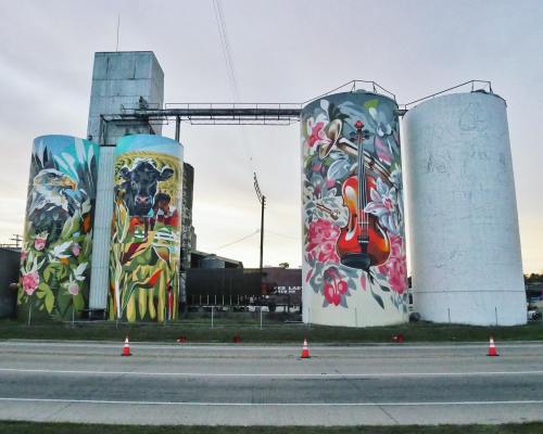 Colorful murals on silos featuring flowers, a dog, and a violin, set against a twilight sky.