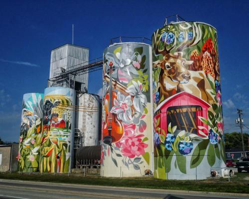 Colorful murals on silos featuring animals, flowers, and a violin against a blue sky.