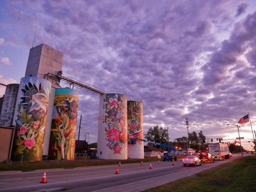 Colorful murals on silos at sunset, with cars on the road and a cloudy sky in the background.