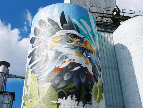 A vibrant mural of an eagle on a cylindrical silo, set against a blue sky with clouds.