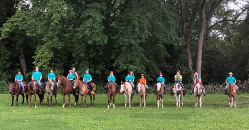 A group of riders in turquoise shirts on horseback, lined up in front of lush green trees.
