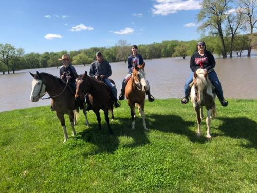 Four people on horseback stand on a grassy area by a river, with trees and blue sky in the background.